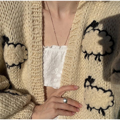 Beige knitted cardigan with sheep patterns worn by a person, showing a hand and ring.