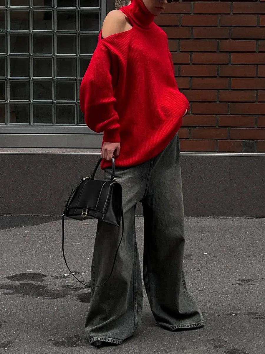 Person wearing a red sweater and gray pants holding a black handbag against a brick wall.