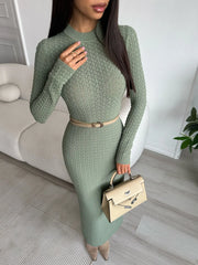 Woman wearing a green knit dress holding a beige handbag indoors.