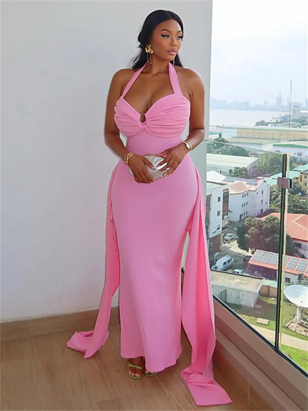 Woman wearing a pink halter neck dress with a high slit, standing by a window with a view of buildings and trees.