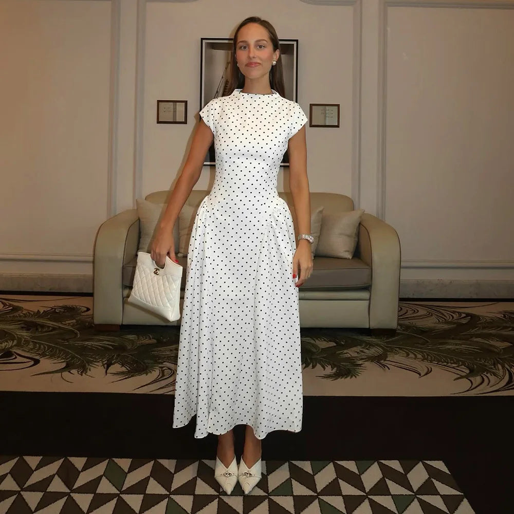 Woman in a white polka dot dress standing in an elegant room with a sofa and framed pictures.