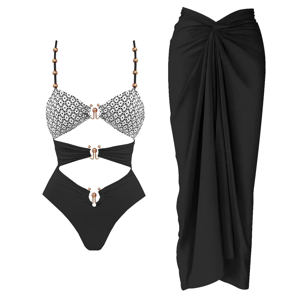 Black and white patterned cutout one-piece swimsuit with embellished straps and matching black draped cover-up skirt.