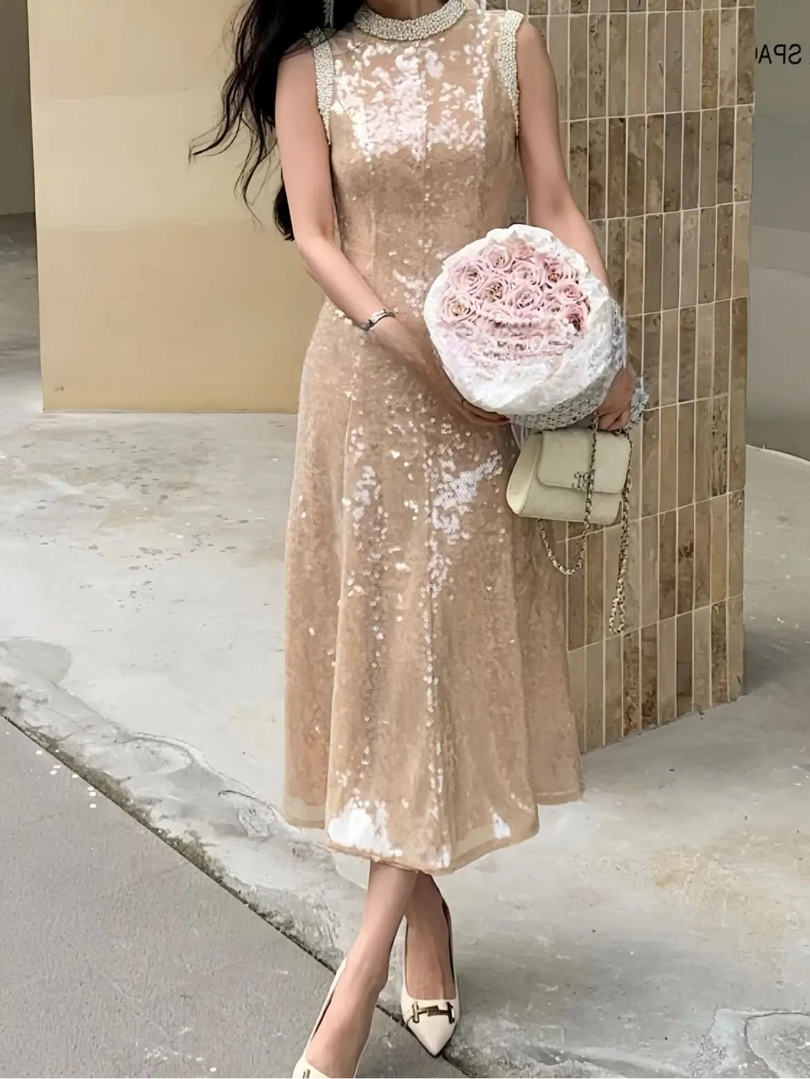 Woman in a beige sequin dress holding flowers and a clutch against a tiled wall.