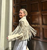 Woman wearing a white fringed jacket standing in front of a wooden door.