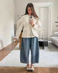 Woman taking a mirror selfie in a stylish outfit with a white blazer and blue pants in a modern living room.