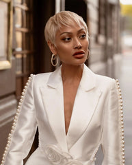 Woman in a white blazer with a deep neckline and pearl details, standing in an elegant interior setting.