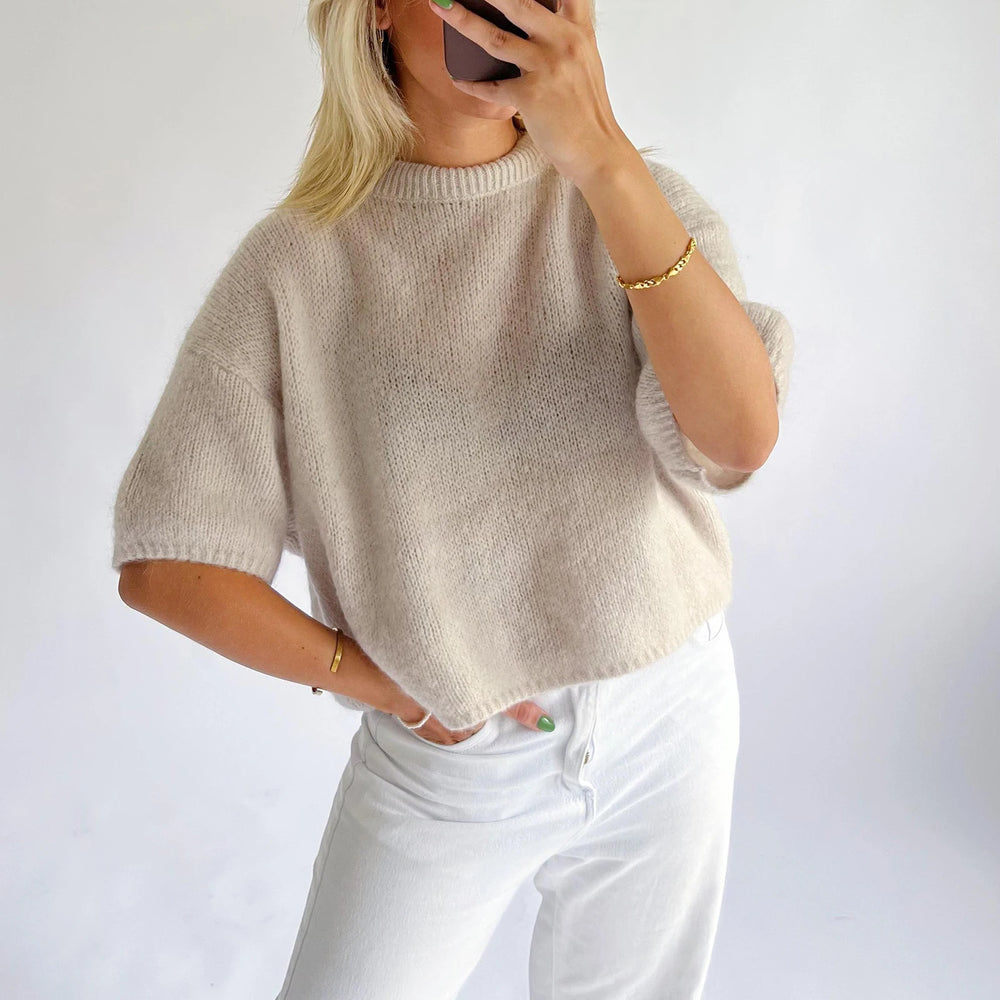 Person wearing a beige knit sweater and white pants against a plain background