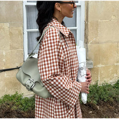 Woman in a red and white checkered dress holding a green bag and a white paper roll, standing against a stone building.