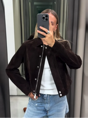Person wearing a brown jacket with black buttons, white shirt, and blue jeans taking a mirror selfie.