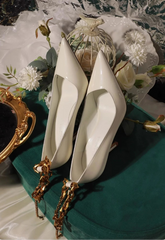 White high-heeled shoes with gold straps on a decorative table with flowers and a teacup.