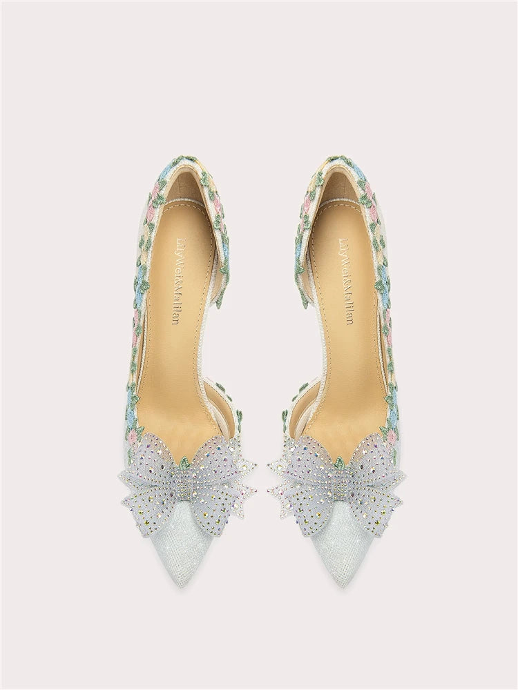 Pair of high-heeled shoes with floral straps and decorative bows on a light gray background