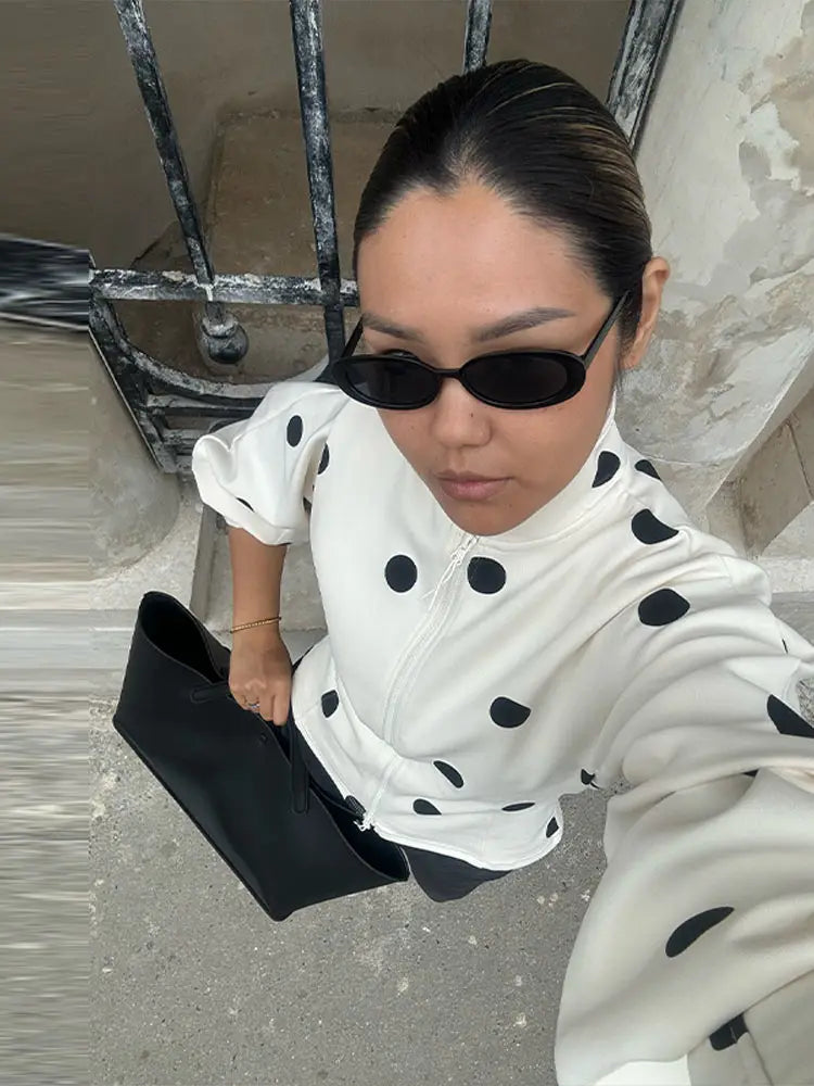 Person wearing a white outfit with black polka dots and sunglasses, holding a black clutch.