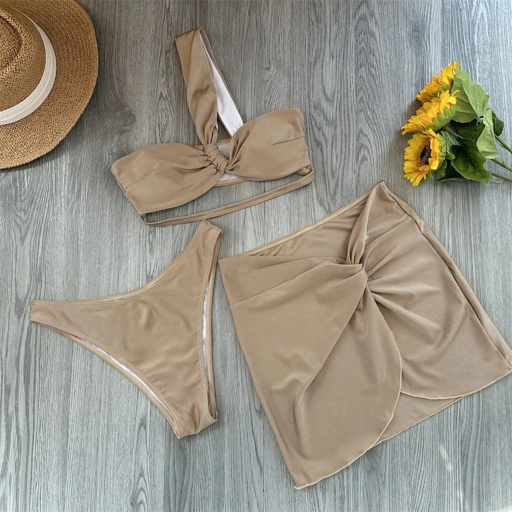 Flat lay of a beige twisted one shoulder bikini top, high-cut bottoms, and a knotted wrap skirt with a straw hat and sunflowers.