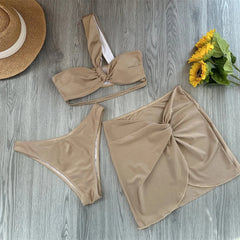 Flat lay of a beige twisted one shoulder bikini top, high-cut bottoms, and a knotted wrap skirt with a straw hat and sunflowers.