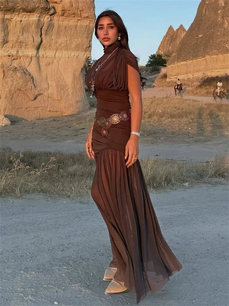 Woman in a brown dress standing in a desert landscape with rock formations.