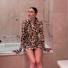 Woman in a leopard print dress sitting on the edge of a bathtub in a tiled bathroom.
