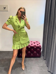 Woman in a green outfit taking a mirror selfie in a room with a purple tufted ottoman.