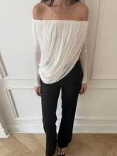 Person wearing a white off-shoulder top and black pants against a white wall.