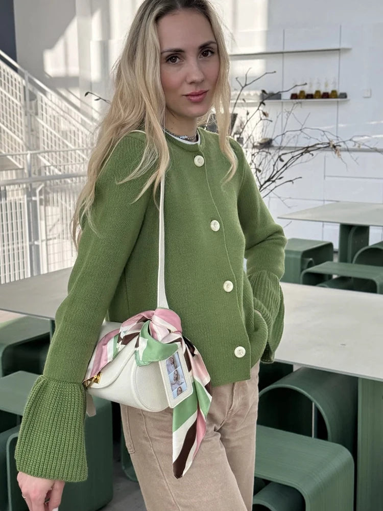 Woman in a green sweater with ruffled sleeves holding a white handbag with a colorful scarf in an indoor setting.