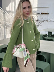 Woman in a green sweater with ruffled sleeves holding a white handbag with a colorful scarf in an indoor setting.