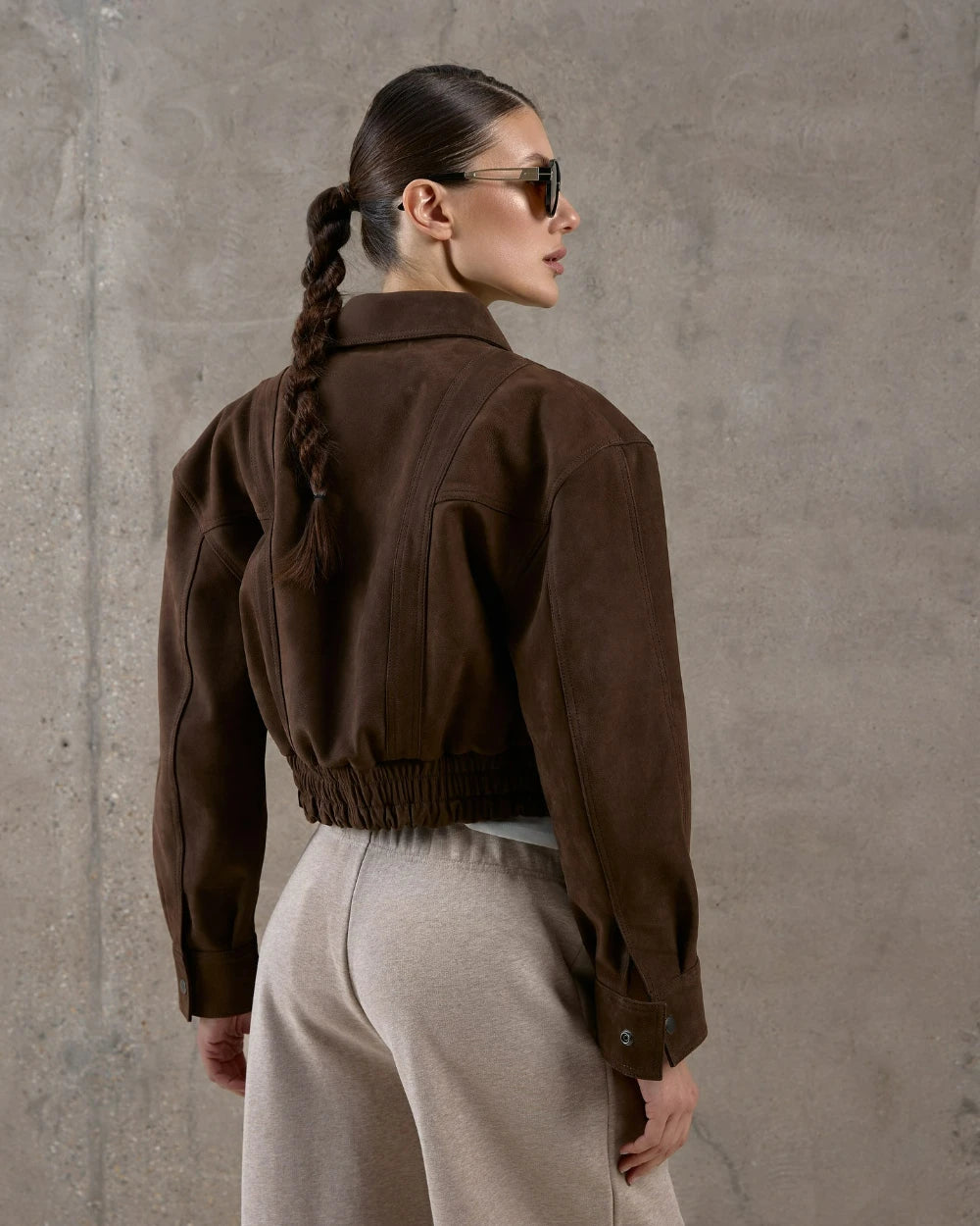 Woman wearing a brown jacket and sunglasses against a gray concrete wall