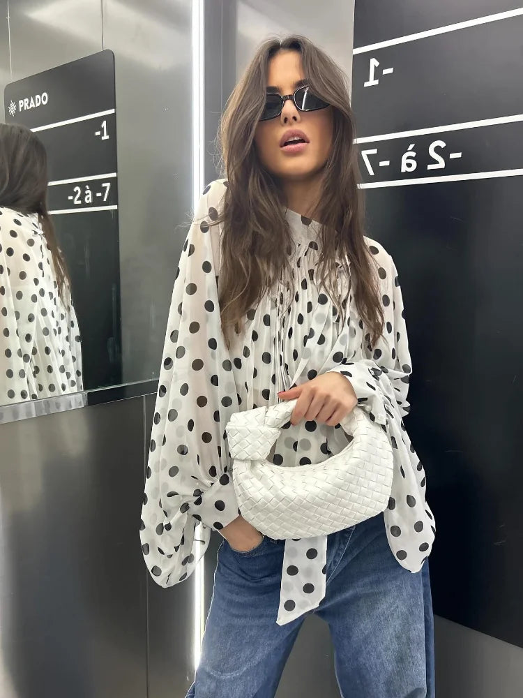 Woman in a polka dot blouse and jeans holding a white bag in front of a mirror.