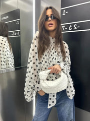 Woman in a polka dot blouse and jeans holding a white bag in front of a mirror.