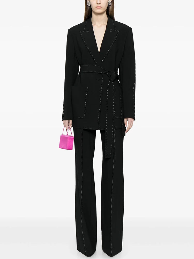 Black belted blazer and wide-leg pants suit with contrast stitching worn by a model, showcasing the front view and a pink mini handbag.