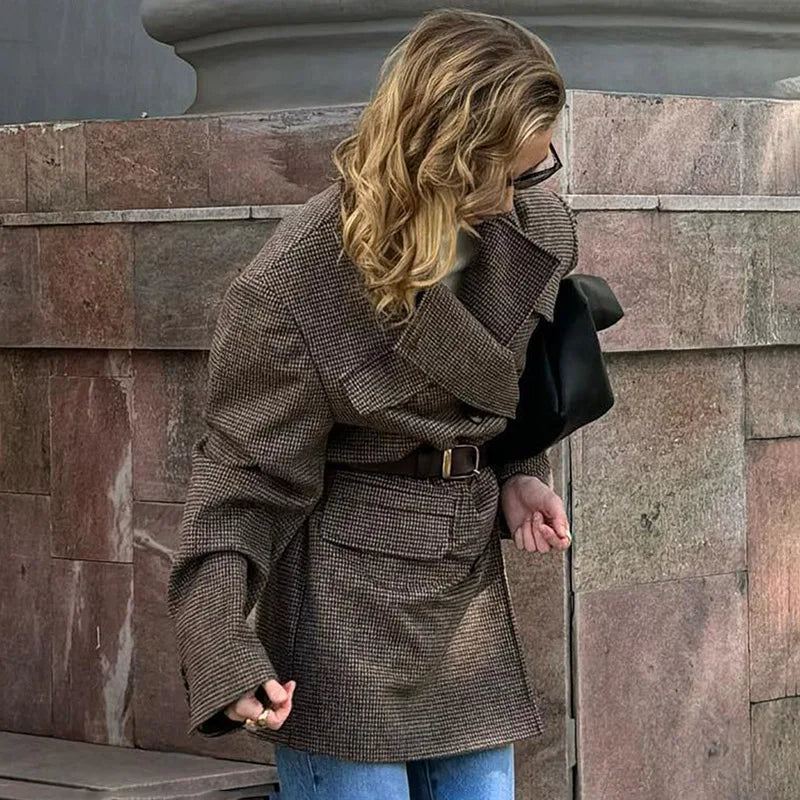 Woman wearing a brown coat standing against a stone wall.