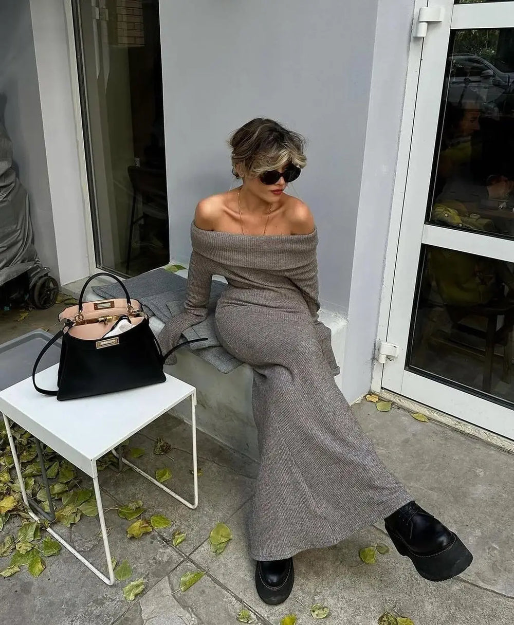 Woman in gray off-shoulder outfit sitting on a chair with a black handbag next to her.
