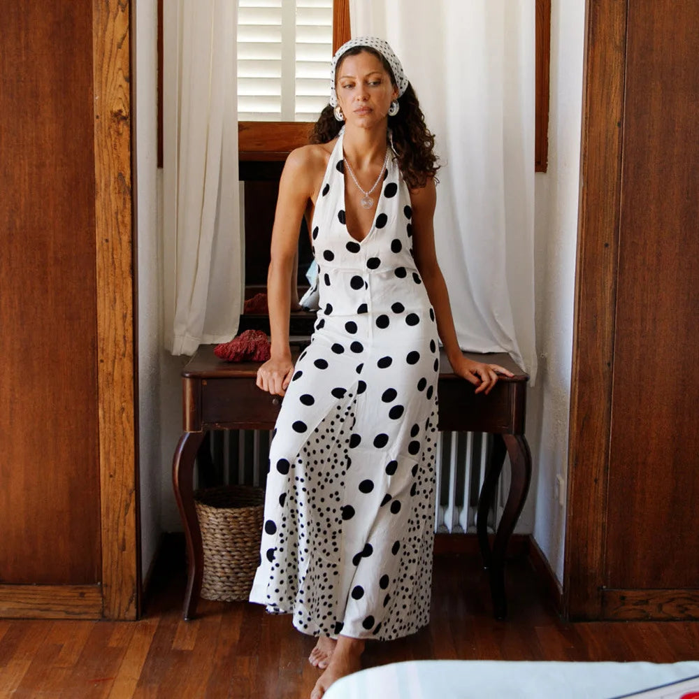 Woman in a white dress with black polka dots sitting on a wooden stool in a room with wooden walls and white curtains.