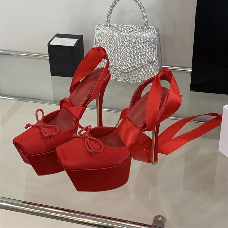 Red high-heeled shoes with ribbon straps on a reflective surface, with a silver handbag in the background.