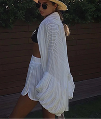Woman in a white outfit with a hat and sunglasses standing against a wooden fence.
