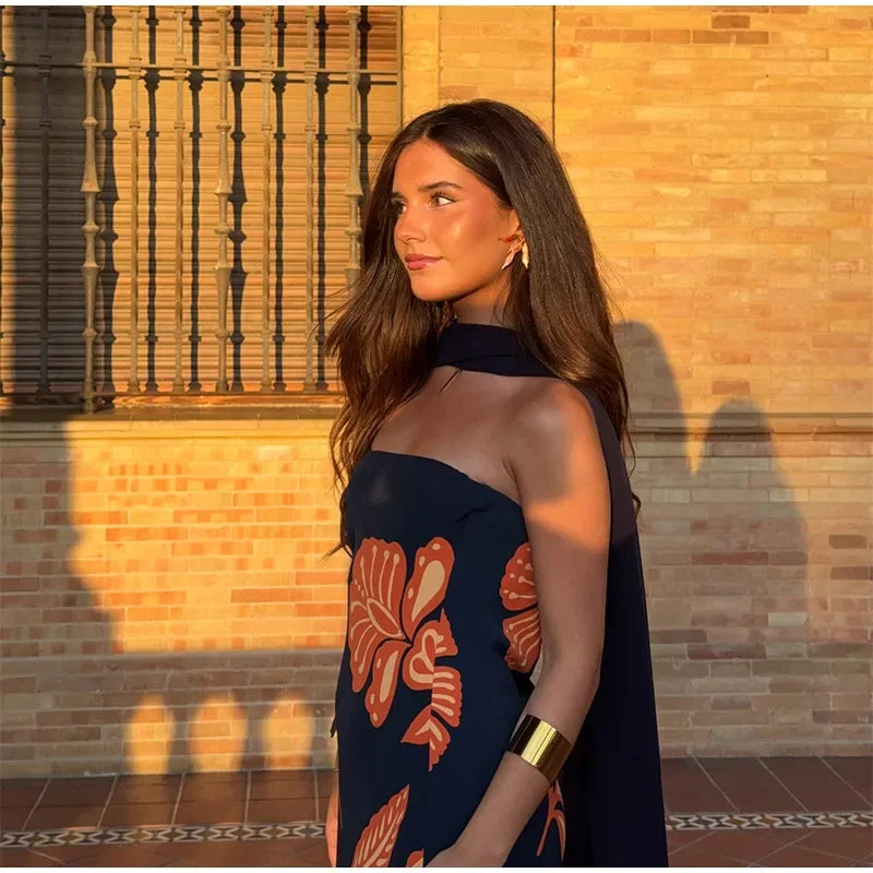 Woman in a dark blue dress with red floral patterns standing against a brick wall.
