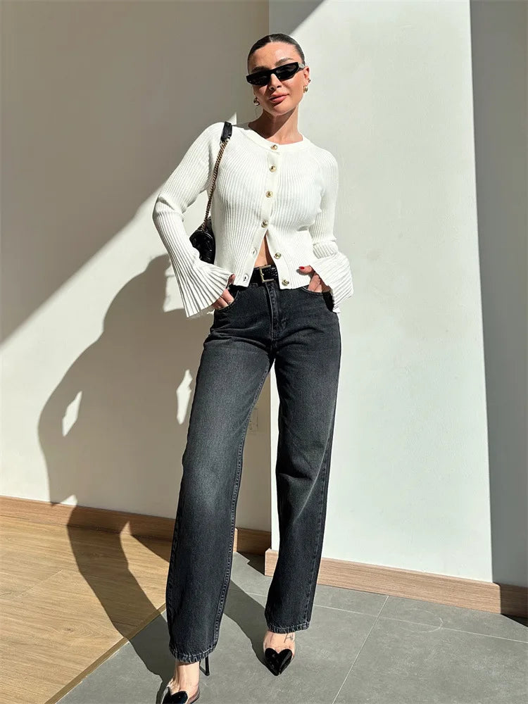 Woman wearing a white cardigan and black jeans standing against a light-colored wall.