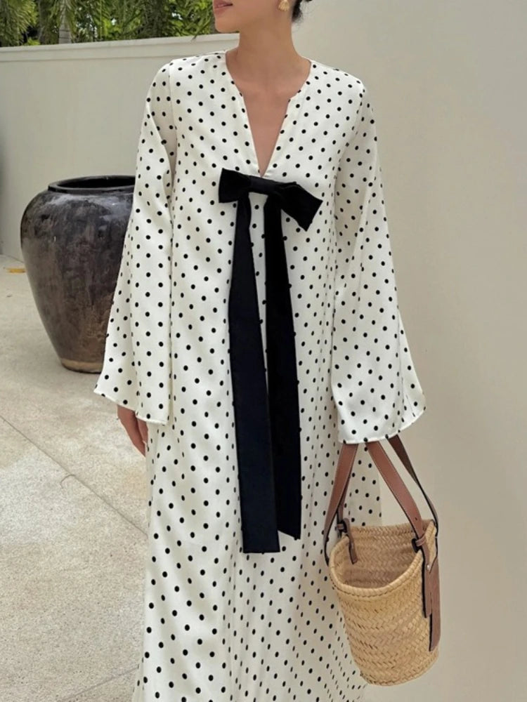 Person wearing a polka dot dress with a black bow, holding a woven bag outdoors.