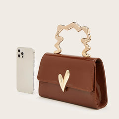 Brown patent leather evening bag with sculpted gold heart handle next to a smartphone.