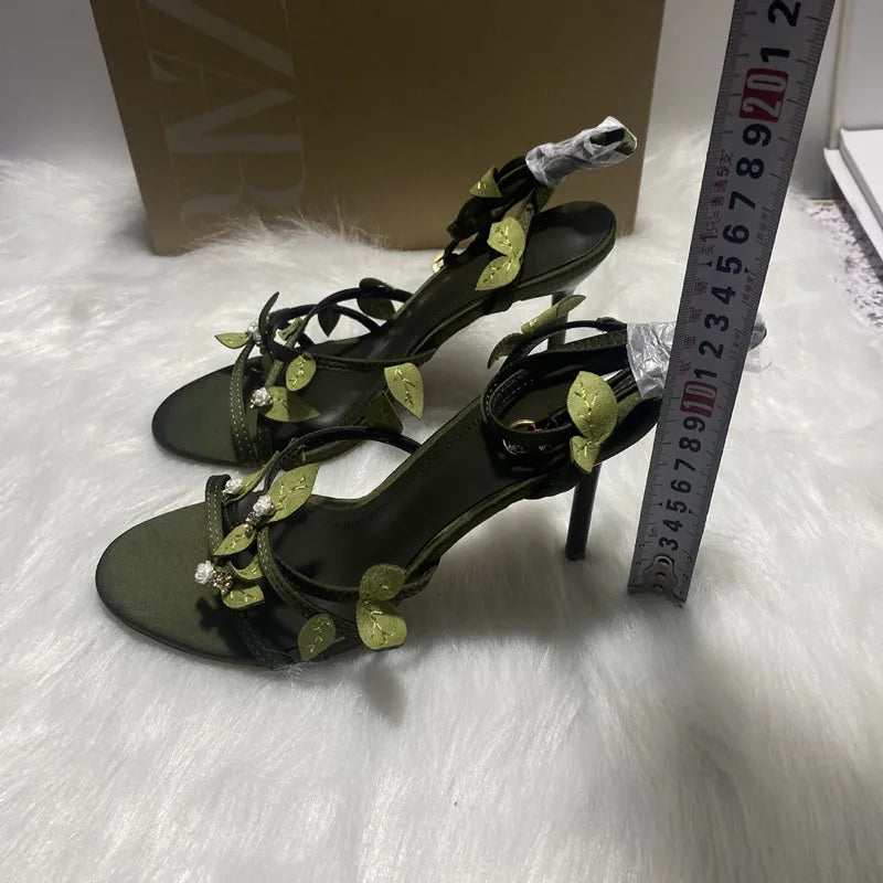 Black high-heeled sandals with green embellishments next to a measuring tape on a white surface.