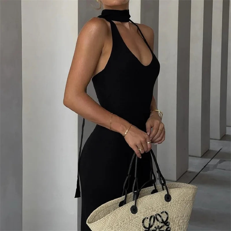 Woman in a black dress holding a straw bag with a brand logo against a striped wall.