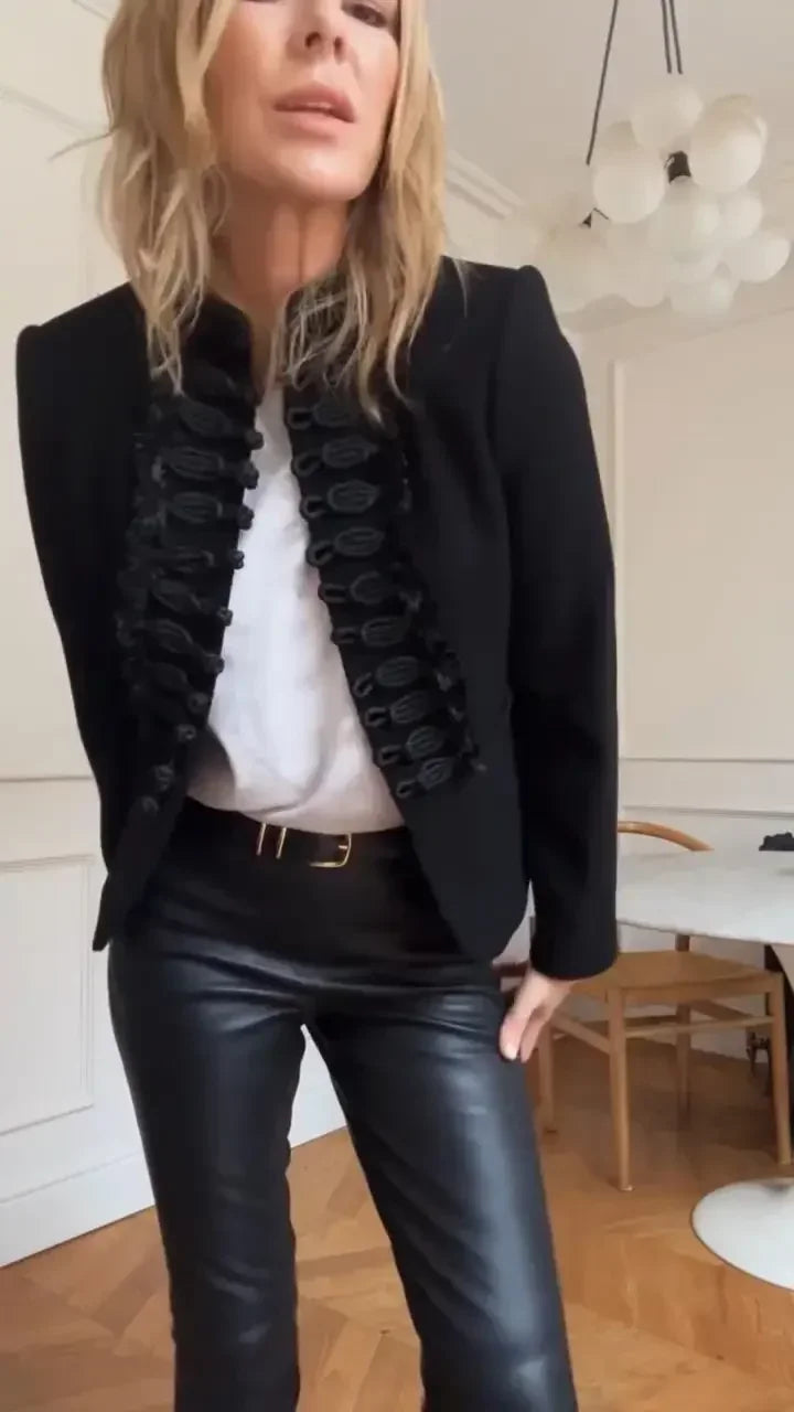 Person wearing a black blazer with ruffled details, white shirt, and black leather pants indoors.