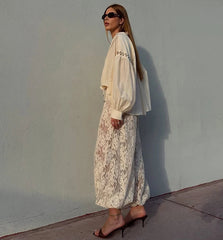 Woman in a light-colored outfit standing against a plain wall.