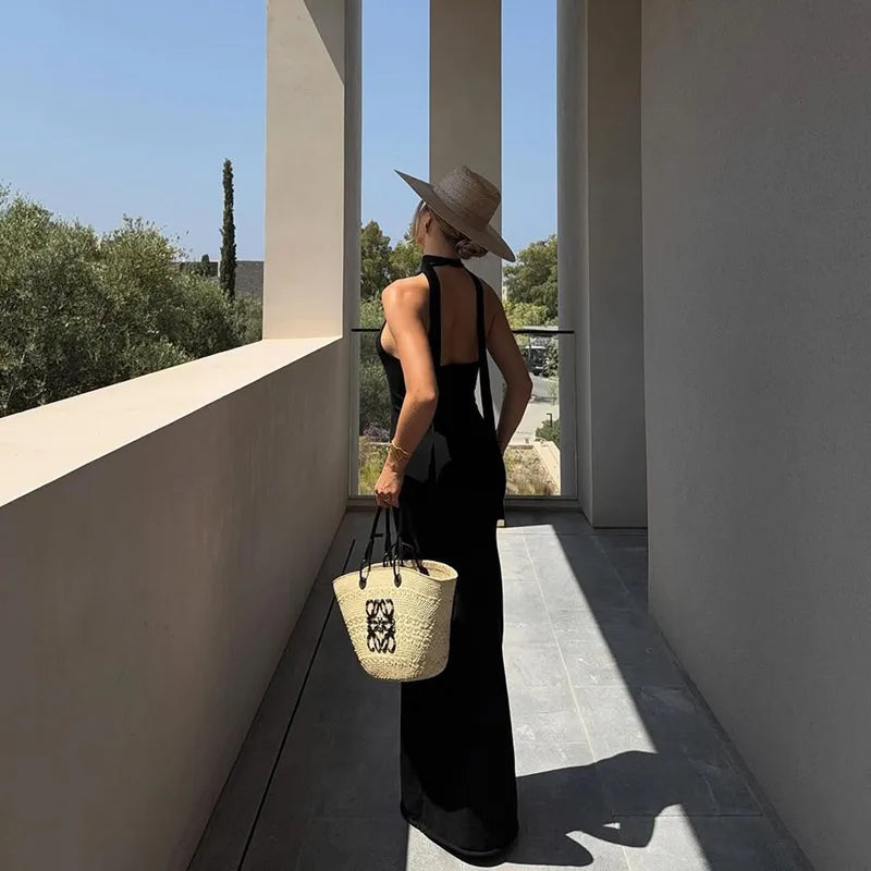 Woman in a black dress and straw hat holding a woven bag on a balcony.