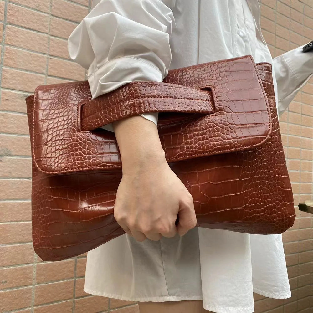 Brown crocodile-patterned clutch held by a person against a brick wall.