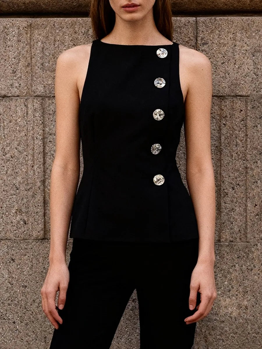 Person wearing a black sleeveless top with silver buttons against a stone wall.
