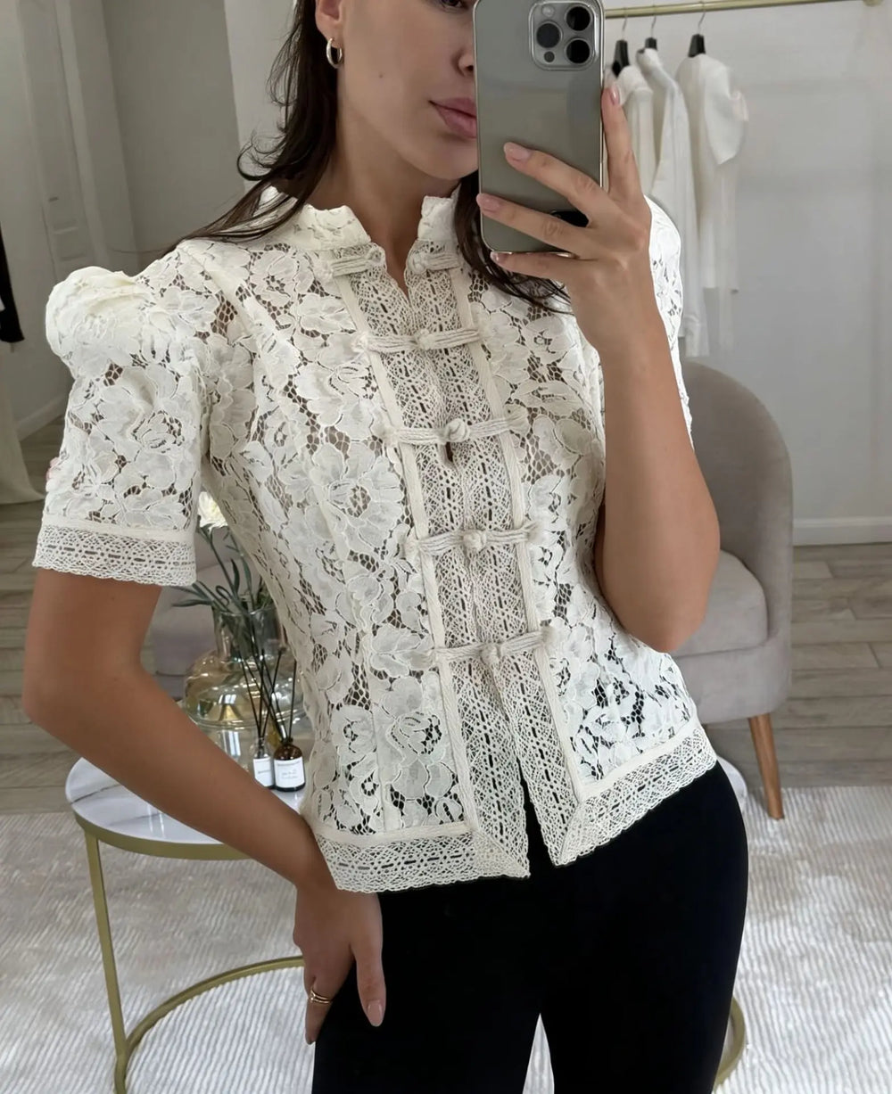 Woman taking a mirror selfie wearing a white lace blouse in a room.