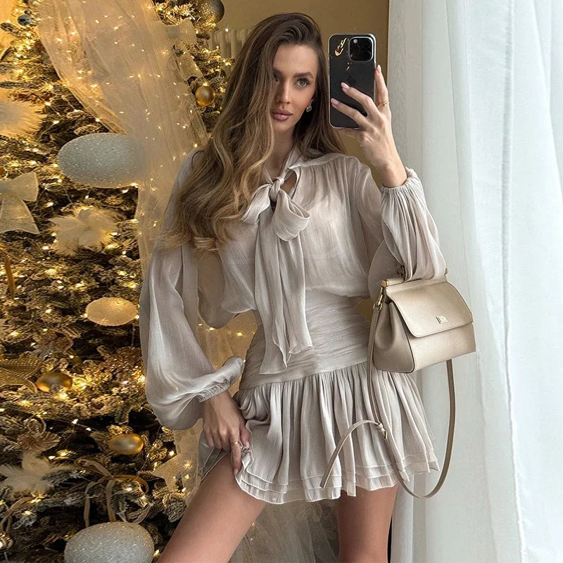 Woman taking a mirror selfie in front of a decorated Christmas tree, wearing a white dress and holding a beige handbag.
