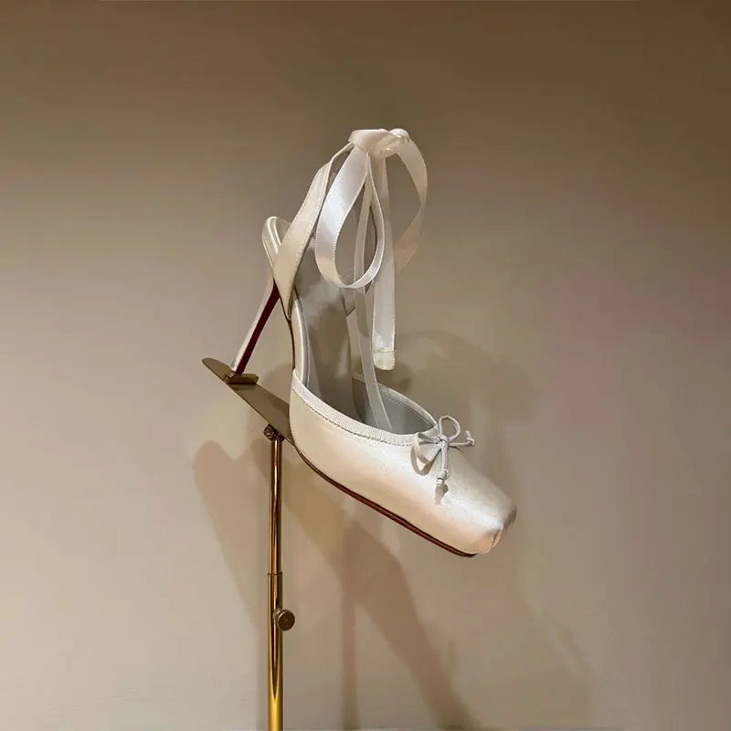 White high-heeled shoes with ribbon details on a stand against a beige background