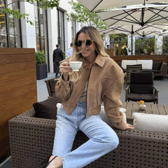A woman sitting outdoors holding a cup, wearing sunglasses and a khaki jacket.