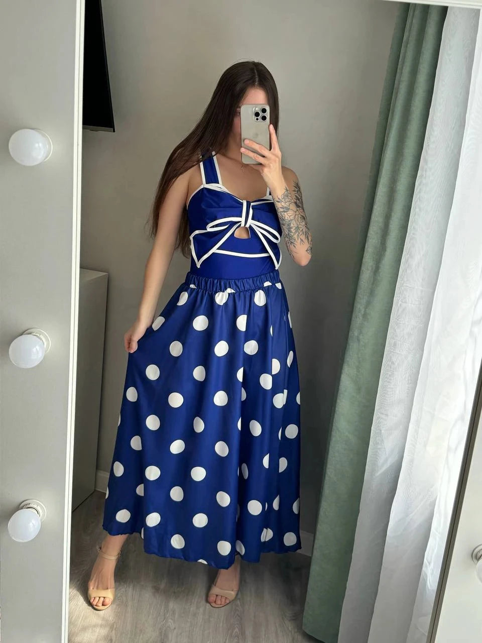 Woman wearing a navy blue polka dot tankini swimsuit with a white bow detail and matching skirt, posing for a mirror selfie.