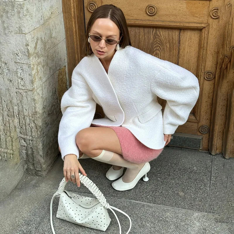 Woman in a white coat and pink skirt holding a white handbag, crouching on a street.